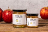 Two jars of Laurel Hill Apple Jelly on a rustic wooden table, with fresh red apples in the background against a textured light gray wall.