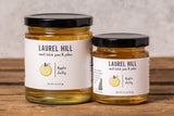 Two glass jars of Laurel Hill Apple Jelly with black lids and white labels, displayed on a rustic wooden table against a textured light gray wall.