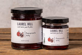 Two jars of Laurel Hill Pomegranate Jelly on a rustic wooden table against a textured light gray wall.