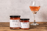 Two jars of Laurel Hill Rosé Wine Jelly with black lids and white labels, displayed on a rustic wooden table next to a glass of iced root beer against a textured light gray wall.