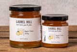 Two glass jars of Laurel Hill Apple Cider Jelly on a rustic wooden table against a textured light gray wall.