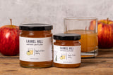Two jars of Laurel Hill Apple Cider Jam on a rustic wooden table, styled with fresh red apples and a glass of the amber-colored jam against a textured light gray wall.