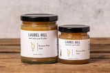 Two glass jars of Laurel Hill banana rum jam on a rustic wooden table against a textured light gray wall.