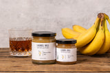 Two jars of Laurel Hill Banana Rum Jam shown on a rustic wooden table, with a glass of amber liquid and a bunch of yellow bananas as props against a light gray textured wall.