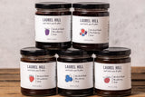 Five jars of Laurel Hill Sweet and Baked Food jam arranged in a pyramid on a rustic wooden surface against a textured light gray wall.