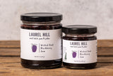 Two jars of Laurel Hill Blackberry Jam on a rustic wooden table against a textured light gray wall.