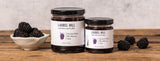 Two jars of Laurel Hill Blackberry Jam on a rustic wooden table, styled with fresh blackberries and a small bowl of blackberries against a textured light gray background.