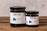 Two jars of Laurel Hill Small Batch Blueberry Jam on a rustic wooden surface against a textured light gray wall.