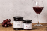 Two jars of Laurel Hill Cabernet Wine Jelly on a rustic wooden table, styled with a glass of red wine and a cluster of red grapes against a textured light gray background.