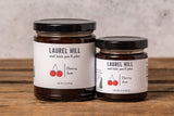 Two jars of Laurel Hill Cherry Jam, one large and one small, standing on a rustic wooden surface against a textured light gray wall.