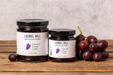 Two jars of Laurel Hill Concord Grape Jam on a rustic wooden table against a textured light gray wall. 
