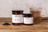 Two glass jars of Laurel Hill tea on a rustic wooden table with a glass mug of brewed tea and loose leaf tea in the background.