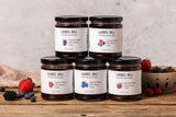 Five jars of Laurel Hill small batch jam featuring blackberry, multiberry, strawberry, blueberry and raspberry flavors on a rustic wood table.