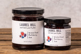 Two jars of Laurel Hill Wicked Good Multiberry Jam on a rustic wooden table against a light gray textured wall. The larger jar is labeled 'Mulberry Jam' and the smaller jar is labeled 'Wild Game Blackberries Jam'.