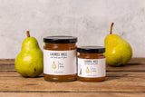 Two jars of Laurel Hill Pear Jelly displayed on a rustic wooden table, flanked by two fresh green pears against a textured light gray wall.