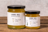 Two glass jars of Laurel Hill Pina Colada Jelly with black lids and white labels, displayed on a rustic wooden surface against a textured light gray wall.