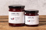 Two glass jars of Laurel Hill jam, one larger Raspberry Lavender Jam jar and one smaller Raspberry Lavender Jam jar, displayed on a rustic wooden surface against a textured light gray wall.