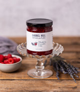 A jar of Laurel Hill Raspberry Lavender Jam displayed on a glass pedestal stand on a rustic wooden table, with a small bowl of fresh raspberries and lavender sprigs in the background.