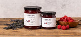 Two jars of Laurel Hill Raspberry Jam on a rustic wooden table, with a small bowl of fresh raspberries and lavender sprigs against a textured light gray background.