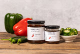 Two jars of Laurel Hill Red Pepper Jam arranged on a rustic wooden table with fresh peppers and a bowl of sliced jalapeños against a light textured wall.