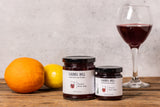 Two jars of Laurel Hill Sangria Jelly on a rustic wooden table, styled with an orange, a lemon, and a glass of red wine against a light gray concrete wall.