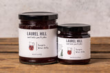 Two glass jars of Laurel Hill Sangria Wine Jelly, one large and one small, sitting on a rustic wooden table against a textured light gray concrete wall.