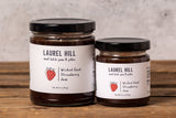 Two glass jars of Laurel Hill Sweetened Fruit Strawberry Jam on a rustic wooden table against a textured light gray wall.